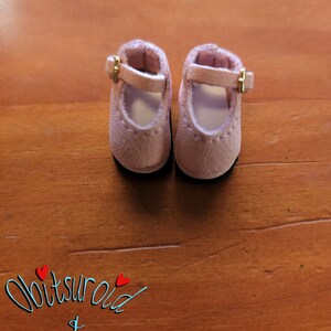 May include: A pair of pink suede doll shoes with gold buckles. The shoes have a simple design and are perfect for a doll's outfit.
