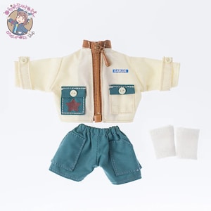 Puede incluir: Un conjunto de ropa para muñeca con una chaqueta de color crema con cremallera, una etiqueta de nombre azul que dice "CARLOS", un pantalón corto verde y un par de calcetines blancos.