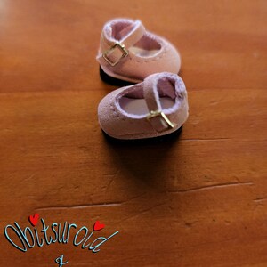 May include: A pair of light pink suede doll shoes with gold buckles. The shoes have a strap across the top and a rounded toe.