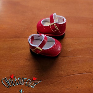 May include: A pair of red doll shoes with white lining and gold buckles. The shoes are on a wooden surface.