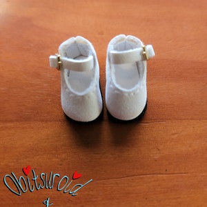 May include: A pair of white doll shoes with gold buckles. The shoes are on a wooden surface.