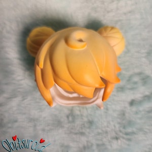 May include: A yellow doll hair wig with two buns on top. The wig is made of a soft, pliable material and is designed to fit a doll's head.