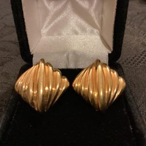 May include: A pair of gold-colored, ridged, square-shaped earrings displayed in a black velvet jewelry box with a white satin interior. The earrings have a textured, shell-like design.