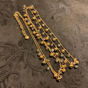 以下が含まれることがあります： 小さな丸い虹色のビーズと装飾的な金色のアクセントが付いた、金色の多層ネックレス。 ネックレスには、留め金にハート型のチャームが付いています。 ジュエリーは、暗く質感のある表面に表示されています。