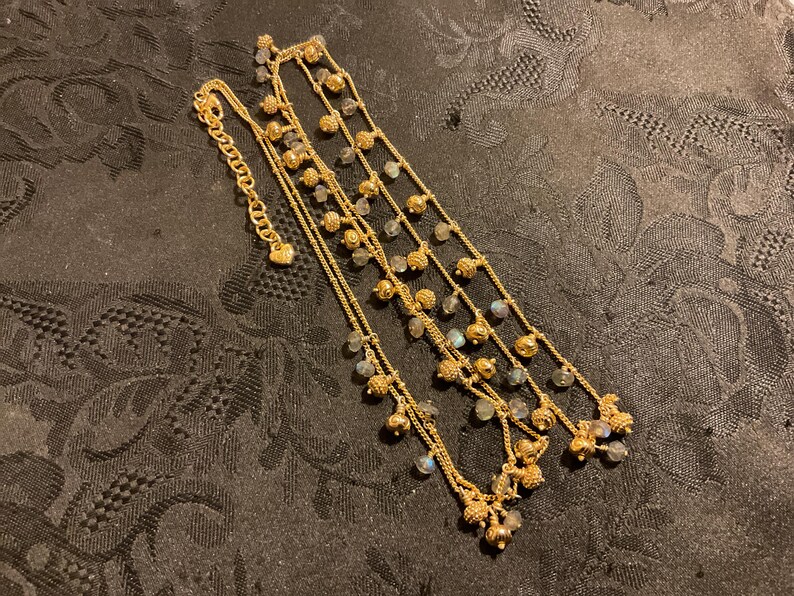 以下が含まれることがあります： 小さな丸い虹色のビーズと装飾的な金色のアクセントが付いた、金色の多層ネックレス。 ネックレスには、留め金にハート型のチャームが付いています。 ジュエリーは、暗く質感のある表面に表示されています。