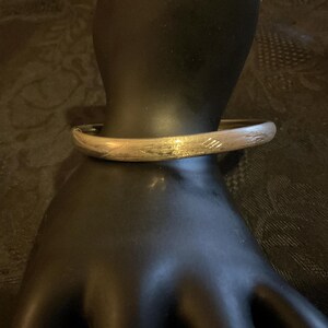 May include: A gold-toned bangle bracelet with etched linear designs. The bracelet is displayed on a black mannequin hand, showcasing its curved shape and metallic sheen. The jewelry piece is a classic accessory.