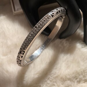 May include: A silver hinged bangle bracelet with a row of small, clear gemstones and decorative scrollwork. The bracelet is displayed on a white, fluffy surface.