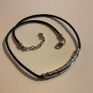 Puede incluir: Collar de cuero trenzado negro con un colgante curvo plateado. El colgante presenta dise&ntilde;os florales y geom&eacute;tricos grabados. El collar tiene un cierre plateado y un extensor de cadena.