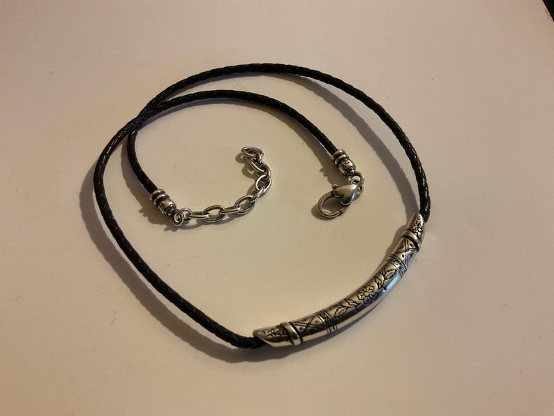 Puede incluir: Collar de cuero trenzado negro con un colgante curvo plateado. El colgante presenta dise&ntilde;os florales y geom&eacute;tricos grabados. El collar tiene un cierre plateado y un extensor de cadena.