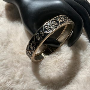 May include: A gold-coloured hinged bangle bracelet with a black enamel inlay. The bracelet features an ornate, scrolling design. The bracelet is resting on a black hand-shaped display against a white, fluffy background.