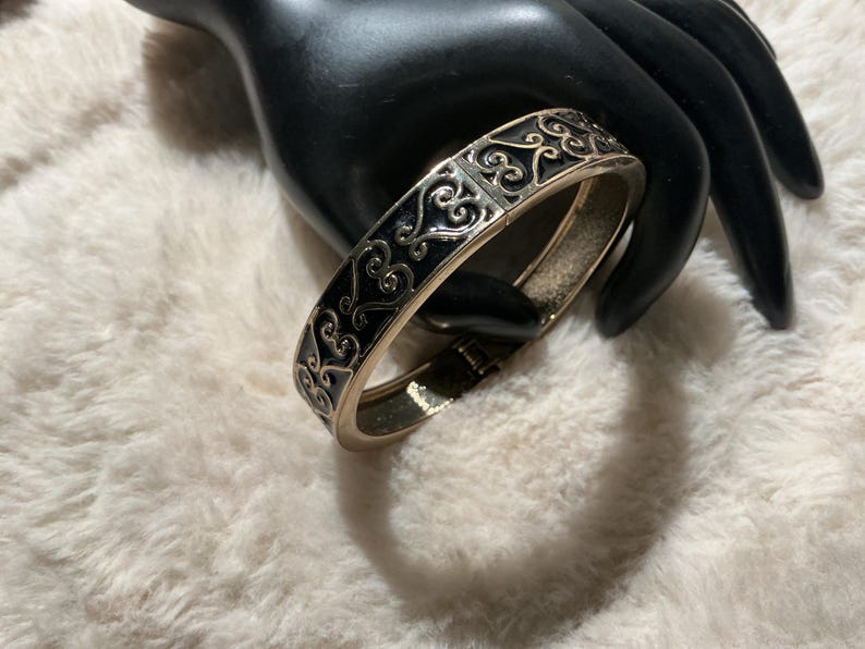 May include: A gold-coloured hinged bangle bracelet with a black enamel inlay. The bracelet features an ornate, scrolling design. The bracelet is resting on a black hand-shaped display against a white, fluffy background.