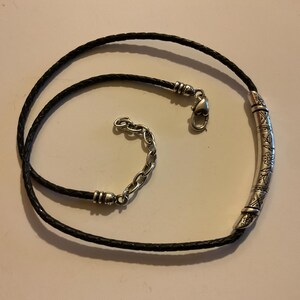 Puede incluir: Un collar de cuero negro con un colgante de barra de metal plateado. El colgante tiene un dise&ntilde;o floral grabado. El collar tiene un cierre de mosquet&oacute;n y una cadena ajustable. El collar est&aacute; sobre una superficie clara.