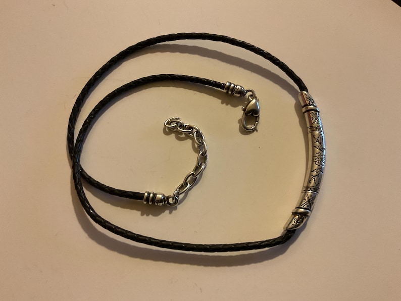 Puede incluir: Un collar de cuero negro con un colgante de barra de metal plateado. El colgante tiene un dise&ntilde;o floral grabado. El collar tiene un cierre de mosquet&oacute;n y una cadena ajustable. El collar est&aacute; sobre una superficie clara.