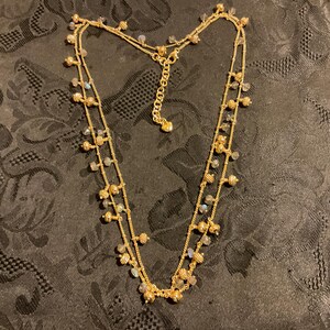 以下が含まれることがあります： 2連のゴールドトーンのネックレス。小さな丸いビーズと虹色の石のクラスターで飾られています。ネックレスは調節可能なチェーン付きで、暗い模様の表面に展示されています。繊細なジュエリーです。