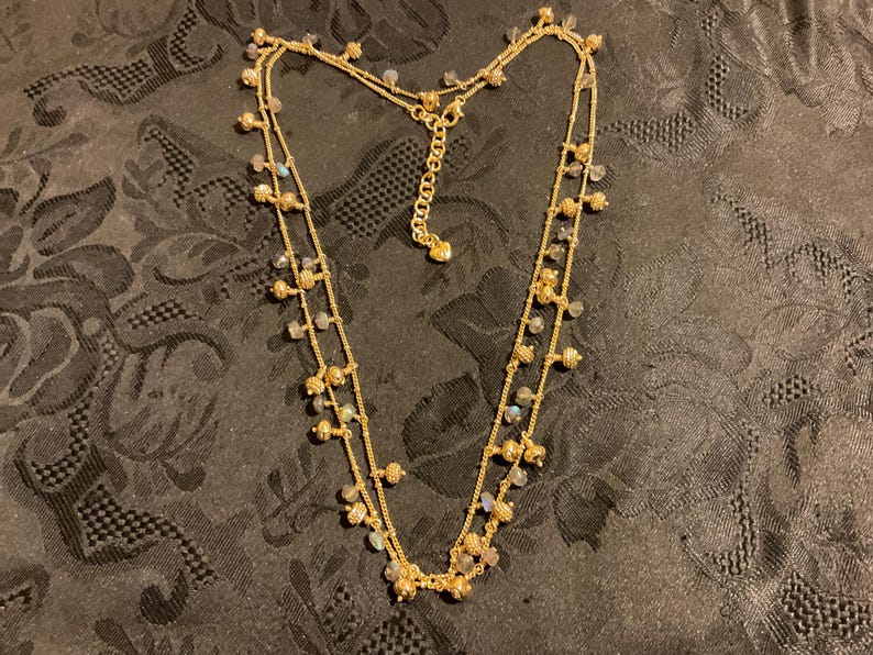 以下が含まれることがあります： 2連のゴールドトーンのネックレス。小さな丸いビーズと虹色の石のクラスターで飾られています。ネックレスは調節可能なチェーン付きで、暗い模様の表面に展示されています。繊細なジュエリーです。