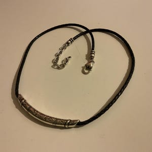 Puede incluir: Collar de cuero trenzado negro con un colgante de barra de metal plateado. El colgante tiene un dise&ntilde;o grabado. El collar tiene un cierre de mosquet&oacute;n y una cadena ajustable.