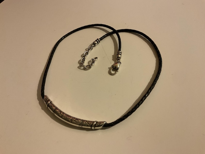 Puede incluir: Collar de cuero trenzado negro con un colgante de barra de metal plateado. El colgante tiene un dise&ntilde;o grabado. El collar tiene un cierre de mosquet&oacute;n y una cadena ajustable.