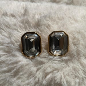 May include: A pair of rectangular, gray-toned crystal stud earrings with gold-colored frames. The earrings are set against a fluffy, white background, creating a contrast between the cool crystals and the warm, textured backdrop.