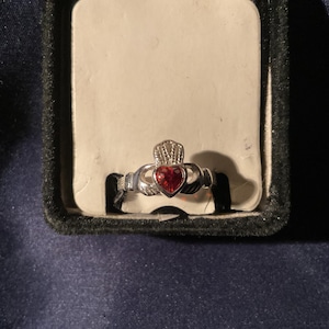 May include: A silver-toned Claddagh ring with a red heart-shaped stone. The ring features a crown above the heart, held by two hands. The ring is displayed in a black jewelry box with a white insert.