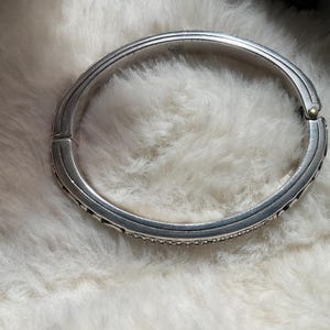 May include: A silver hinged bangle bracelet with a textured finish. The bracelet features small, sparkling details along the edges. The clasp is visible, and the bracelet is set against a soft, white background.