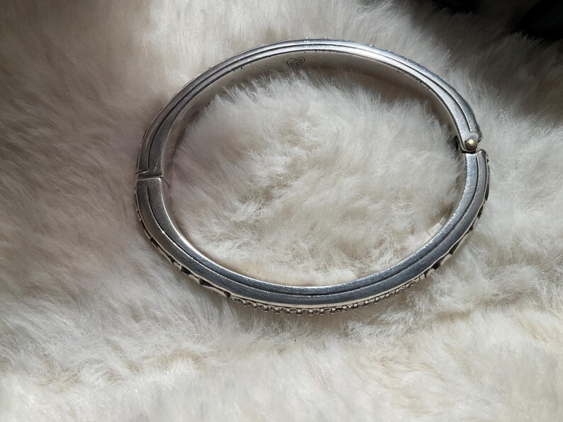 May include: A silver hinged bangle bracelet with a textured finish. The bracelet features small, sparkling details along the edges. The clasp is visible, and the bracelet is set against a soft, white background.