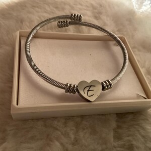 May include: A silver-toned bracelet with a heart-shaped charm. The charm features the letter "E" in a delicate script. The bracelet is presented in a white box, resting on a soft, textured surface.
