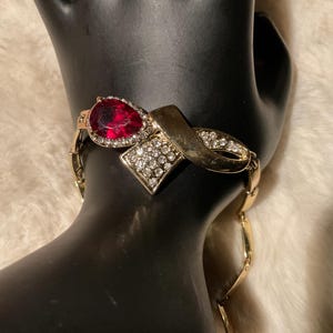 May include: Gold-toned bracelet featuring a large, pear-shaped, red gemstone and a diamond-studded accent. The bracelet has a ribbon-like design with additional small, clear gemstones. The bracelet is displayed on a black mannequin hand.