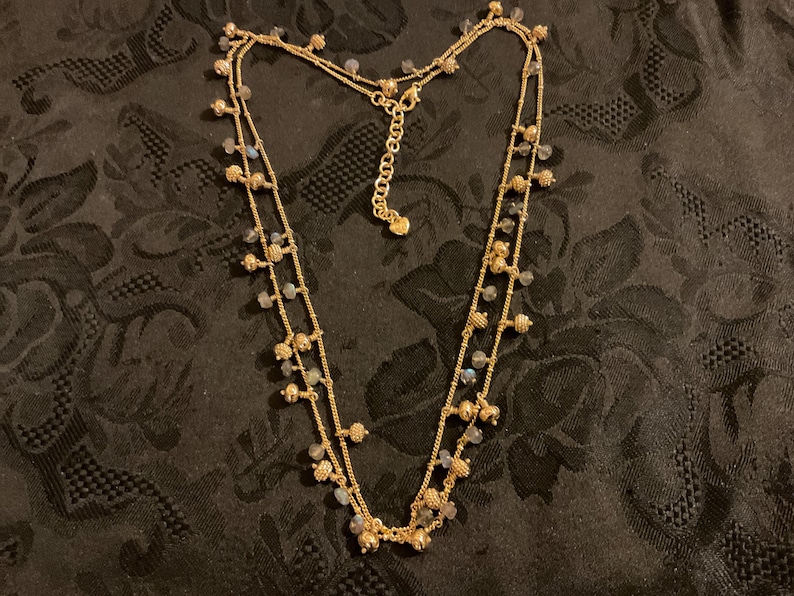 以下が含まれることがあります： 2連のゴールドトーンネックレス。小さな丸いビーズと、小さなイリデッセントビーズのクラスターで飾られています。ネックレスは調節可能なチェーンと繊細でフェミニンなデザインで、様々な場面に適しています。