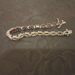 May include: A silver bracelet featuring oval-cut, deep red gemstones. The bracelet is composed of individual links, each holding a gemstone. The clasp is visible, and the bracelet is displayed on a dark, textured surface.