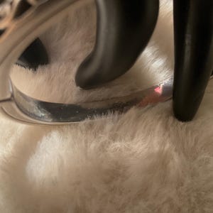 May include: A silver bangle bracelet with a decorative pattern. The bracelet is resting on a white, fluffy surface. The bracelet has a curved design and appears to be made of metal.