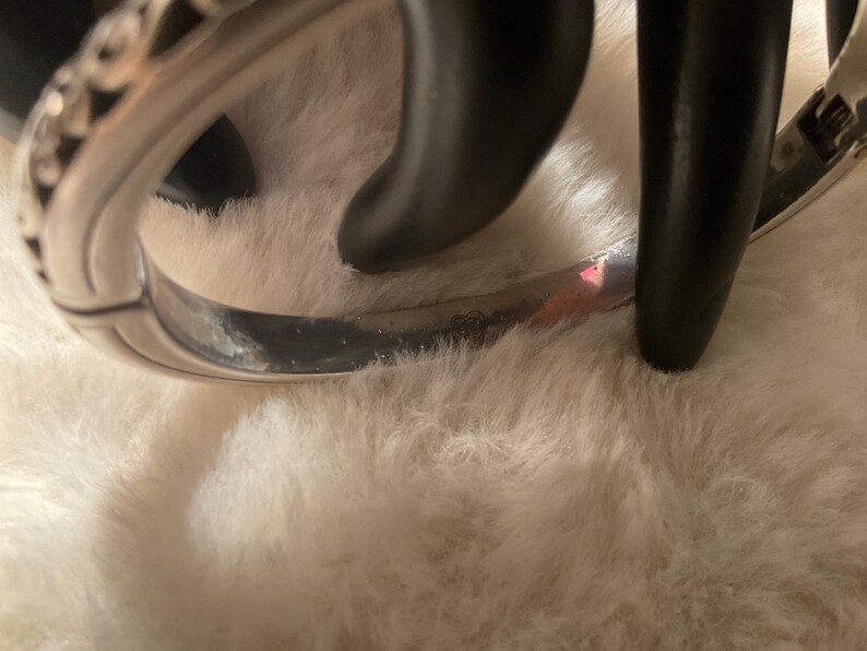 May include: A silver bangle bracelet with a decorative pattern. The bracelet is resting on a white, fluffy surface. The bracelet has a curved design and appears to be made of metal.