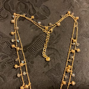 以下が含まれることがあります： 複数のストランドを持つゴールドトーンのネックレス。ネックレスは、小さく丸い金のビーズと、小さく、薄い青と透明なビーズのアクセントが特徴です。ネックレスは、暗い模様のある表面に表示されています。