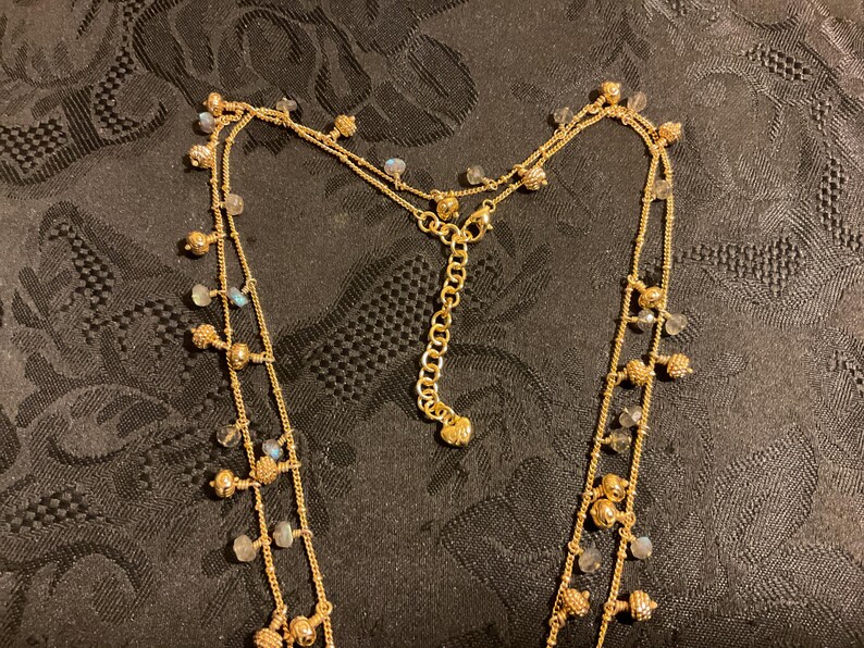 以下が含まれることがあります： 複数のストランドを持つゴールドトーンのネックレス。ネックレスは、小さく丸い金のビーズと、小さく、薄い青と透明なビーズのアクセントが特徴です。ネックレスは、暗い模様のある表面に表示されています。