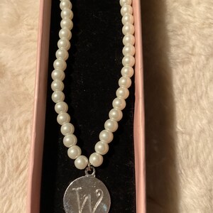 May include: A pearl necklace with a silver pendant featuring the letter "W" inside a pink box. The necklace is made of round, white pearls. The pendant is circular and has a shiny finish.