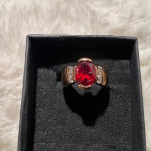 May include: A gold ring with a large, oval-cut red gemstone as the centerpiece. The ring has smaller, clear gemstones on each side of the main stone. The ring is set in a black jewelry box.