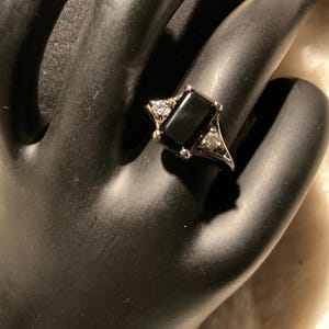 May include: A silver ring featuring a rectangular black stone as the centerpiece, flanked by smaller, clear stones. The ring is set on a black surface, showcasing its design and details.