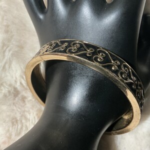 May include: Gold-coloured hinged bangle bracelet with a black enamel inlay. The bracelet features an ornate, scrolling design. The bracelet is shown on a black hand-shaped display against a soft, white background.