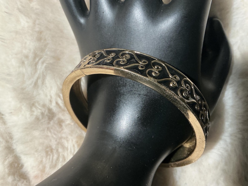 May include: Gold-coloured hinged bangle bracelet with a black enamel inlay. The bracelet features an ornate, scrolling design. The bracelet is shown on a black hand-shaped display against a soft, white background.