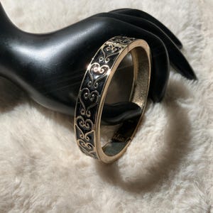 May include: A gold-coloured hinged bracelet with a black enamel inlay featuring a repeating heart and scroll design. The bracelet is displayed on a black hand-shaped jewelry holder against a white, fluffy background.