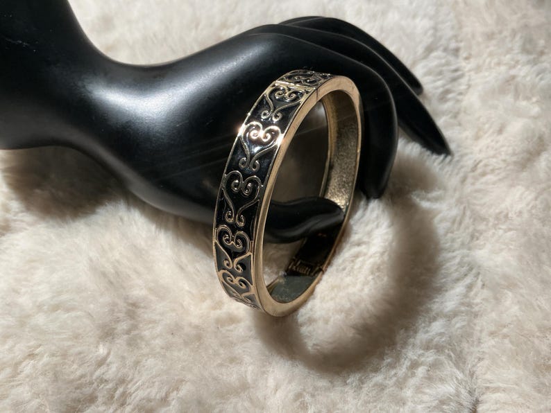 May include: A gold-coloured hinged bracelet with a black enamel inlay featuring a repeating heart and scroll design. The bracelet is displayed on a black hand-shaped jewelry holder against a white, fluffy background.