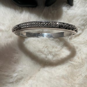May include: Silver-coloured hinged bangle bracelet. The top is encrusted with small, clear crystals. The sides feature an ornate, swirling design. The bracelet is resting on a white, fluffy surface.
