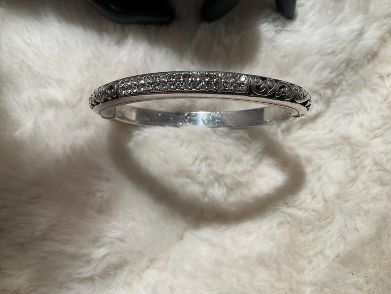 May include: Silver-coloured hinged bangle bracelet. The top is encrusted with small, clear crystals. The sides feature an ornate, swirling design. The bracelet is resting on a white, fluffy surface.