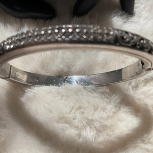 May include: A silver hinged bangle bracelet with detailed embellishments. The bracelet has a decorative pattern along the top edge and a smooth interior. The clasp is visible, and the bracelet appears to be made of metal.
