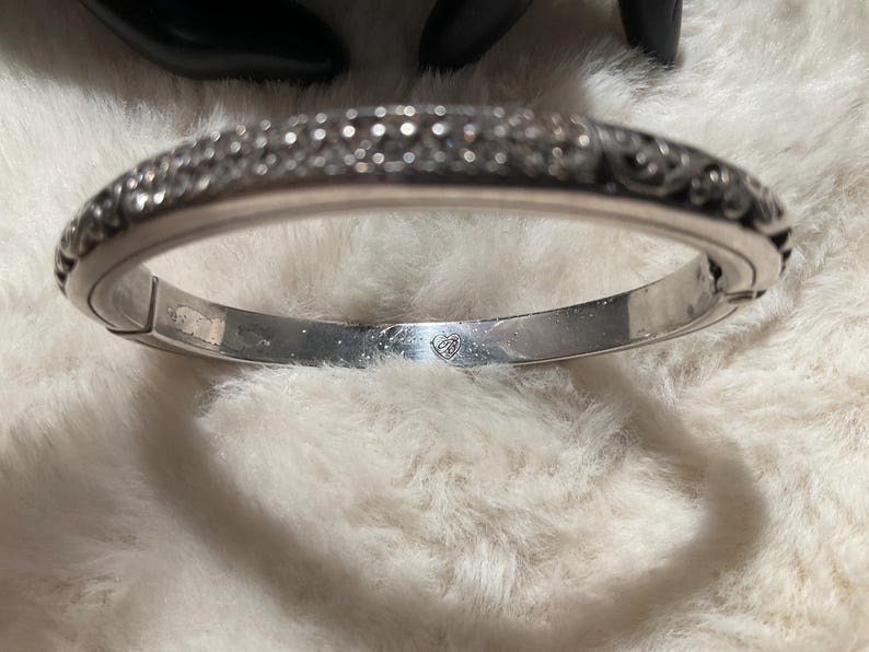 May include: A silver hinged bangle bracelet with detailed embellishments. The bracelet has a decorative pattern along the top edge and a smooth interior. The clasp is visible, and the bracelet appears to be made of metal.
