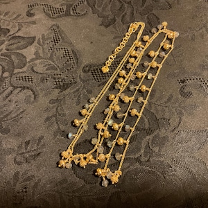 以下が含まれることがあります： 複数のストランドを持つ金色のネックレス。ネックレスは、小さくて丸い金色のビーズと、小さくて明るい色の石のクラスターが特徴です。ネックレスは、暗い模様のある表面に表示されています。