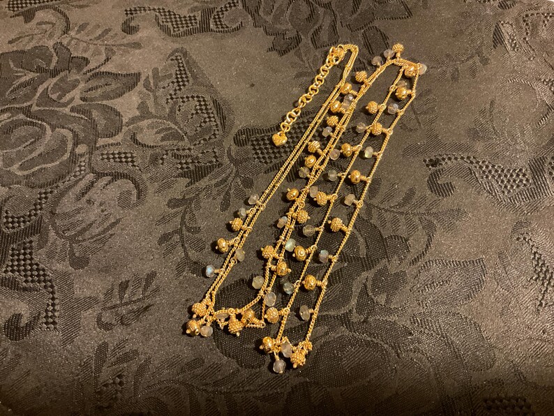 以下が含まれることがあります： 複数のストランドを持つ金色のネックレス。ネックレスは、小さくて丸い金色のビーズと、小さくて明るい色の石のクラスターが特徴です。ネックレスは、暗い模様のある表面に表示されています。