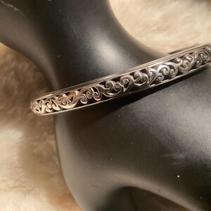 May include: A silver bangle bracelet featuring an elaborate, swirling filigree pattern. The bracelet is presented on a black mannequin arm, highlighting its detailed openwork design. The metal has a polished finish.