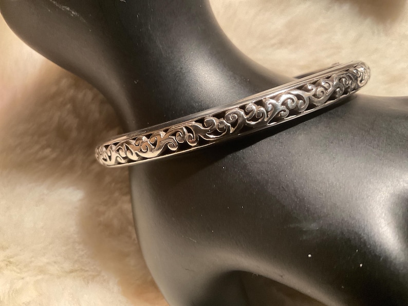 May include: A silver bangle bracelet featuring an elaborate, swirling filigree pattern. The bracelet is presented on a black mannequin arm, highlighting its detailed openwork design. The metal has a polished finish.