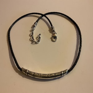 Puede incluir: Un collar de cuero trenzado negro con un colgante de barra de metal plateado. El colgante tiene un dise&ntilde;o grabado. El collar tiene un cierre plateado y una cadena ajustable.