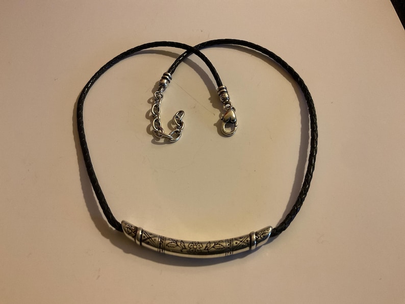 Puede incluir: Un collar de cuero trenzado negro con un colgante de barra de metal plateado. El colgante tiene un dise&ntilde;o grabado. El collar tiene un cierre plateado y una cadena ajustable.